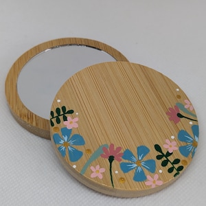 May include: A round bamboo compact mirror with a floral design. The lid is decorated with blue and pink flowers, green leaves, and gold accents. The mirror is open, showing the reflective surface.