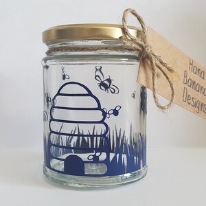 May include: A glass jar with a gold lid and a blue beehive design. The jar is decorated with bees and grass. The jar has a brown paper tag with the text "Hana Banana Designs".