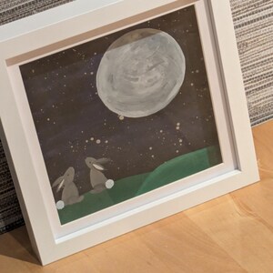 May include: Framed artwork depicting a night scene with a large, grey moon and a starry sky. Two grey rabbits sit on a green hill. The artwork is in a white frame, resting on a wooden surface.