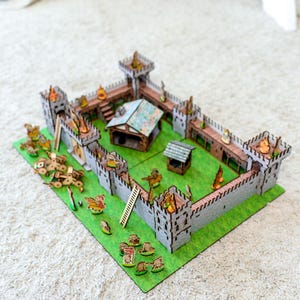 Może przedstawiać: Miniaturowy drewniany model zamku z szarymi murami, brązowymi akcentami i zieloną podstawą. Model zawiera wieże, mały domek, studnię i miniaturowe figurki. Zamek umieszczony jest na zielonej filcowej podstawie.