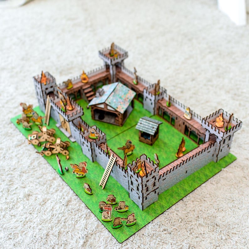 Może przedstawiać: Miniaturowy drewniany model zamku z szarymi murami, brązowymi akcentami i zieloną podstawą. Model zawiera wieże, mały domek, studnię i miniaturowe figurki. Zamek umieszczony jest na zielonej filcowej podstawie.