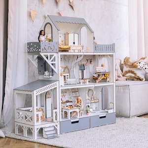 Può includere: Una casa delle bambole in legno a più livelli con tetto grigio e pareti bianche. La casa delle bambole presenta più stanze con mobili e accessori in miniatura. Sono visibili anche una giraffa di peluche e una bambola.