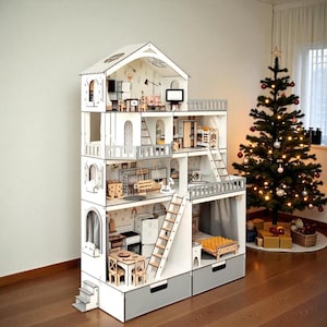 Puede incluir: Una casa de muñecas de madera de varios pisos con un exterior blanco y gris. La casa de muñecas presenta múltiples habitaciones con muebles en miniatura, incluyendo una cocina, sala de estar y dormitorio. Un pequeño árbol de Navidad con adornos está en el fondo.