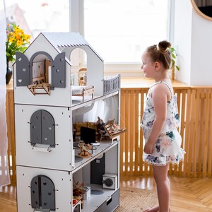 A young girl in a floral dress stands in front of a white and gray wooden dollhouse, looking at the miniature furniture and accessories inside.
