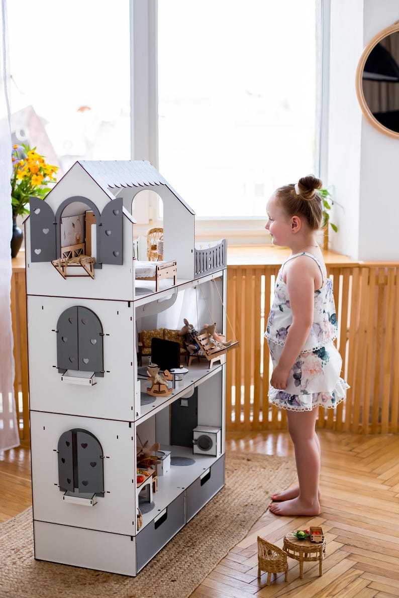 A young girl in a floral dress stands in front of a white and gray wooden dollhouse, looking at the miniature furniture and accessories inside.