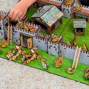 Może przedstawiać: Drewniany zestaw zabawek w postaci zamku z szarymi kamiennymi murami, brązowymi drewnianymi dachami i zieloną podstawą. Zestaw zawiera miniaturowe figurki, drabiny i katapulty. Zamek ma bramę, wieże i mały budynek.