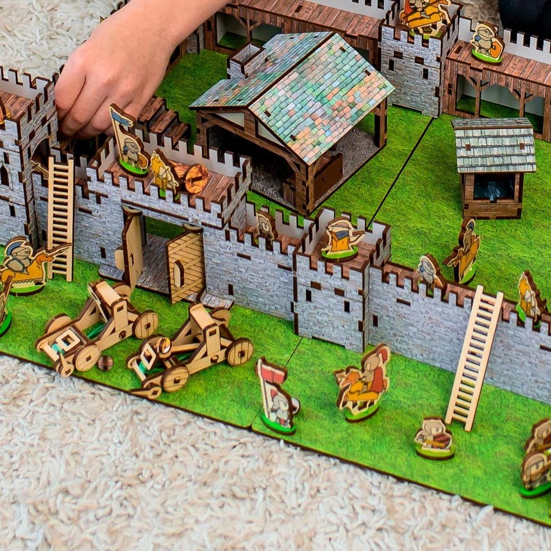 Może przedstawiać: Drewniany zestaw zabawek w postaci zamku z szarymi kamiennymi murami, brązowymi drewnianymi dachami i zieloną podstawą. Zestaw zawiera miniaturowe figurki, drabiny i katapulty. Zamek ma bramę, wieże i mały budynek.