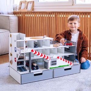 May include: A multi-level wooden toy garage with ramps, parking spaces, and a helipad. The garage is white with gray accents and features various toy cars and trucks. A young child is playing with the toy.