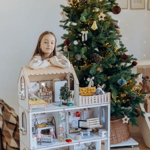 Może przedstawiać: Trzypiętrowy drewniany domek dla lalek z białymi ścianami i wykończeniami z naturalnego drewna. Wnętrze wyposażone jest w miniaturowe meble, w tym łóżka, zestaw kuchenny, stół jadalny, huśtawkę i pralkę oraz drobne elementy dekoracyjne.