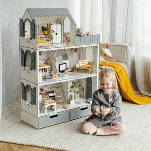 A young girl sits on the floor next to a white and gray wooden dollhouse, holding a toy in her hands.