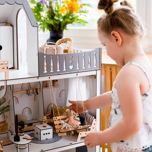 A young girl is playing with a wooden dollhouse, reaching out to interact with the various pieces.
