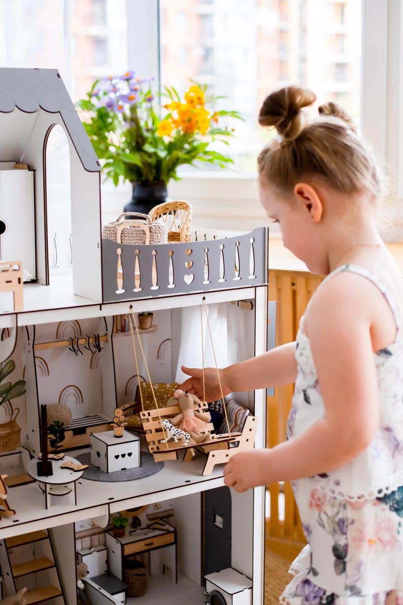 A young girl is playing with a wooden dollhouse, reaching out to interact with the various pieces.