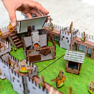 Może przedstawiać: Miniaturowy drewniany zestaw zamkowy z r&oacute;żnymi strukturami, w tym fortecą, studnią i budynkami. Konstrukcje wykonane są z jasnobrązowego drewna i szarego kamienia, ustawione na zielonej powierzchni. Ręka umieszcza dach na jednym z budynk&oacute;w.