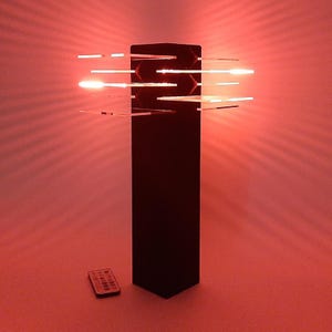May include: A modern black floor lamp with horizontal, illuminated acrylic panels. The lamp emits a warm red glow, with light beams radiating outwards. A small remote control sits on the floor. The lamp's design is sleek and contemporary.