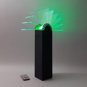 May include: A modern black LED lamp with a rectangular base and a fan-shaped top made of clear, angled panels. The panels are illuminated with green light. A remote control is visible in the foreground.