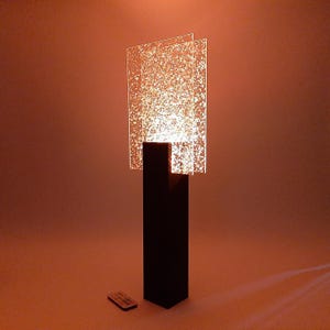 May include: A modern floor lamp with a black rectangular base and a textured, illuminated panel. The panel is composed of three overlapping rectangular glass pieces with a speckled pattern. A remote control sits on the floor.