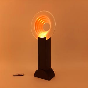 May include: A modern, artistic lamp with a circular, layered design that emits a warm orange glow. The lamp sits on a black rectangular base. A remote control is visible in the foreground. The lamp's design is reminiscent of a spiral.