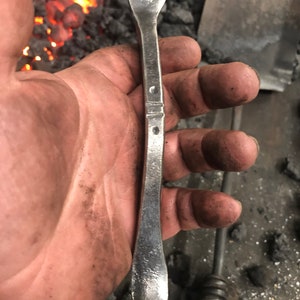 May include: A close-up of a silver metal spoon with a rounded, slightly pointed end and three holes in the top. The spoon is held in a hand, with the background out of focus.