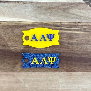 May include: Two decorative tags with the Greek letters ΑΛΨ. One tag is yellow with blue letters, and the other is blue with glitter and yellow letters. The tags are on a wooden cutting board.