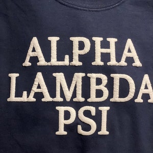 May include: A navy blue t-shirt with white lettering that reads "Alpha Lambda Psi".