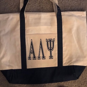 May include: A beige canvas tote bag with black handles and trim. The bag has a large pocket on the front with the letters "ΑΔΨ" printed in black.