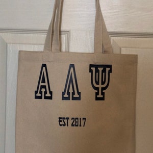 May include: A beige canvas tote bag with black handles and the letters "ΑΑΨ" and the text "EST 2017" printed on the front.
