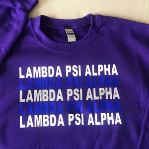 May include: Purple sweatshirt with the text "Lambda Psi Alpha" repeated five times in white letters with a blue glitter effect.