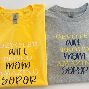 May include: Two t-shirts with the text "Devoted Wife Proud Mom Amazing Soror" printed on them. One shirt is yellow and the other is gray.
