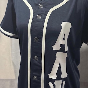 May include: Navy blue baseball jersey with white trim and buttons. The jersey features the Greek letters Alpha, Lambda, and Psi in white on the front. The jersey has short sleeves and a V-neck.