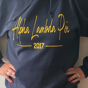 May include: A navy blue hooded sweatshirt with the text "Alpha Lambda Pi" in yellow cursive lettering and "2017" in yellow below.
