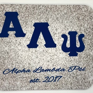 May include: A white mouse pad with a blue and white Greek letter design. The letters are "A". "Λ", and "Ψ". The text "Alpha Lambda Psi est. 2017" is printed below the letters.