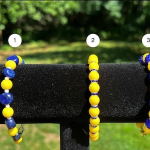 May include: Three bracelets with yellow and blue beads. The first bracelet has smooth blue and yellow beads. The second bracelet has smooth yellow beads and blue beads with white accents. The third bracelet has blue beads with white accents and yellow beads.