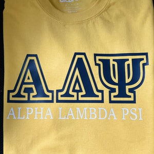 May include: Yellow t-shirt with a navy blue "Alpha Lambda Psi" design. The letters are outlined in black.