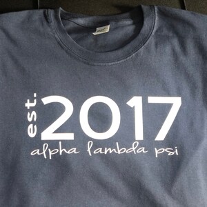 May include: A navy blue t-shirt with white text that reads "est. 2017 alpha lambda psi".