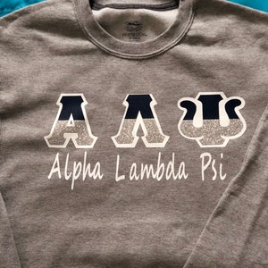 May include: A gray sweatshirt with the letters "A A Ψ" in white with black and silver glitter accents. The words "Alpha Lambda Psi" are written in white below the letters.