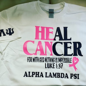 May include: White t-shirt with the words "HEAL CANCER" in pink and black lettering. The text "FOR WITH GOD NOTHING IS IMPOSSIBLE LUKE 1:37" is printed in black below the main text. A pink ribbon with a heart is printed to the right of the text. The words "ALPHA LAMBDA PSI" are printed in black at the bottom of the shirt. The letters "ΑΛΨ" are printed in black on the left sleeve of the shirt.