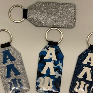 May include: Four keychains with different designs. One keychain is silver glitter, one is gray with blue letters, one is blue camouflage with white letters, and one is blue with white letters. All keychains have a silver ring and a black strap.