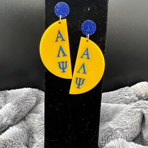 Puede incluir: Un par de pendientes de medio círculo amarillo y azul con las letras griegas Alfa, Lambda y Psi. Los pendientes tienen detalles de purpurina azul.