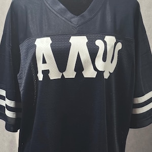 May include: Navy blue mesh jersey with white lettering and stripes. The jersey features a V-neck and white stripes on the sleeves. The Greek letters "AΛΨ" are prominently displayed across the chest in white.