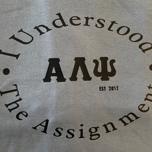 May include: A light blue t-shirt with a black circle graphic. The graphic says "I understood the assignment" with the letters "A". "L", and "Ψ" in a larger font in the center. The text "EST 2017" is below the letters.