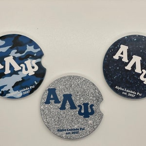 May include: Three round coasters with a blue and white camouflage pattern, a silver glitter pattern, and a dark blue with white stars pattern. Each coaster features the letters "ΑΛΨ" in white, blue, and white respectively. The text "Alpha Lambda Psi est. 2017" is printed on each coaster.