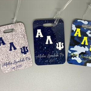 May include: Three luggage tags with different designs. The first tag has a white glitter background with navy blue letters that say "A A Ψ Alpha Lambda Psi est 2017". The second tag has a navy blue glitter background with white letters that say "A A Ψ Alpha Lambda Psi est 2017". The third tag has a blue, white, and black camouflage pattern with yellow letters that say "A A Ψ Alpha Lambda Psi est 2017".