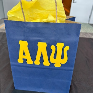 May include: A navy blue paper gift bag with the Greek letters ΑΛΨ in yellow. The bag has brown paper handles and is filled with yellow tissue paper.