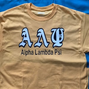 May include: Mustard yellow t-shirt with the Greek letters AΛΨ in white with a black outline. Below the letters is the text "Alpha Lambda Psi" in black. The shirt is made of a soft material.
