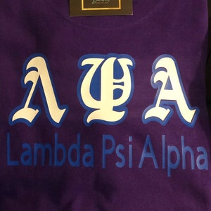 May include: A purple t-shirt with white and blue Greek letters and the words "Lambda Psi Alpha" printed on the front. A small business card is on the top of the shirt.