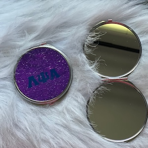 May include: A purple compact mirror with a silver frame and a sparkly finish. The mirror has the name "Anya" printed in blue letters on the front.