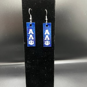 May include: A pair of rectangular earrings with a blue glitter background and white Greek letters. The earrings are suspended from silver-colored hooks. The Greek letters are arranged vertically on each earring.