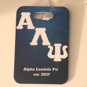 May include: Blue luggage tag with white letters spelling "A A Ψ" and the text "Alpha Lambda Psi est. 2017".