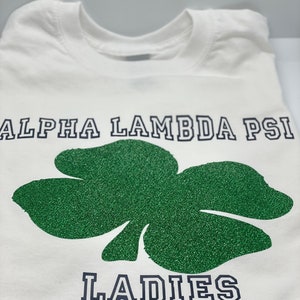 May include: White t-shirt with a green glitter shamrock and the text "ALPHA LAMBDA PSI LADIES" in navy blue.