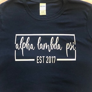 May include: A dark blue t-shirt with white text that reads "alpha lambda psi" in a rectangular box with the words "EST 2017" below the box.
